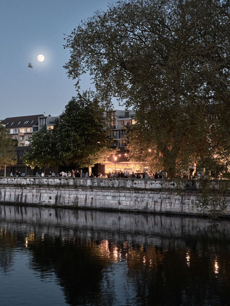 Architekturfotografie: Menschen sitzen und stehen bei der Buvette volAare am Kreuzackerplatz in Solothurn, einer temporären modularen Gastronomie am Aareufer.