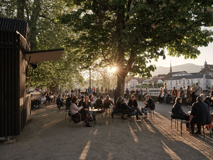 Architekturfotografie: Menschen sitzen und stehen bei der Buvette volAare am Kreuzackerplatz in Solothurn, einer temporären modularen Gastronomie am Aareufer.