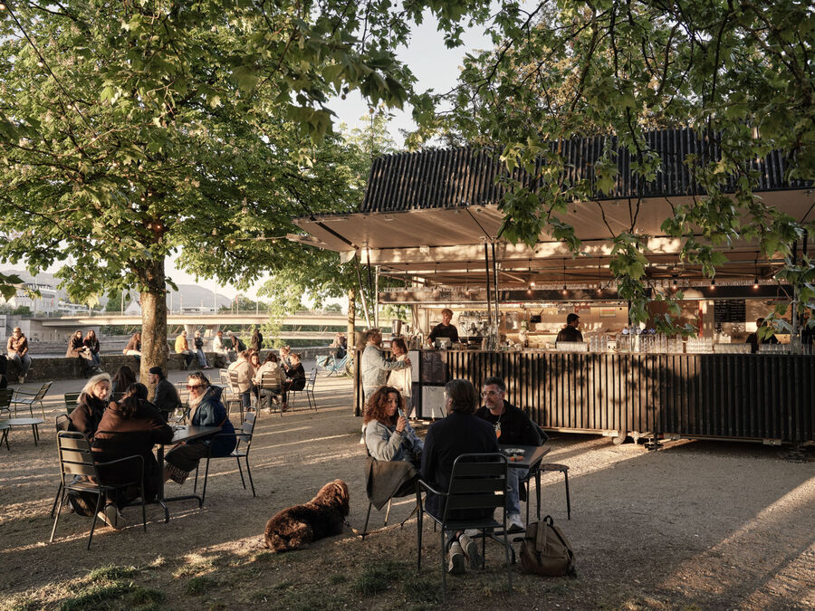 Architekturfotografie: Menschen sitzen und stehen bei der Buvette volAare am Kreuzackerplatz in Solothurn, einer temporären modularen Gastronomie am Aareufer.