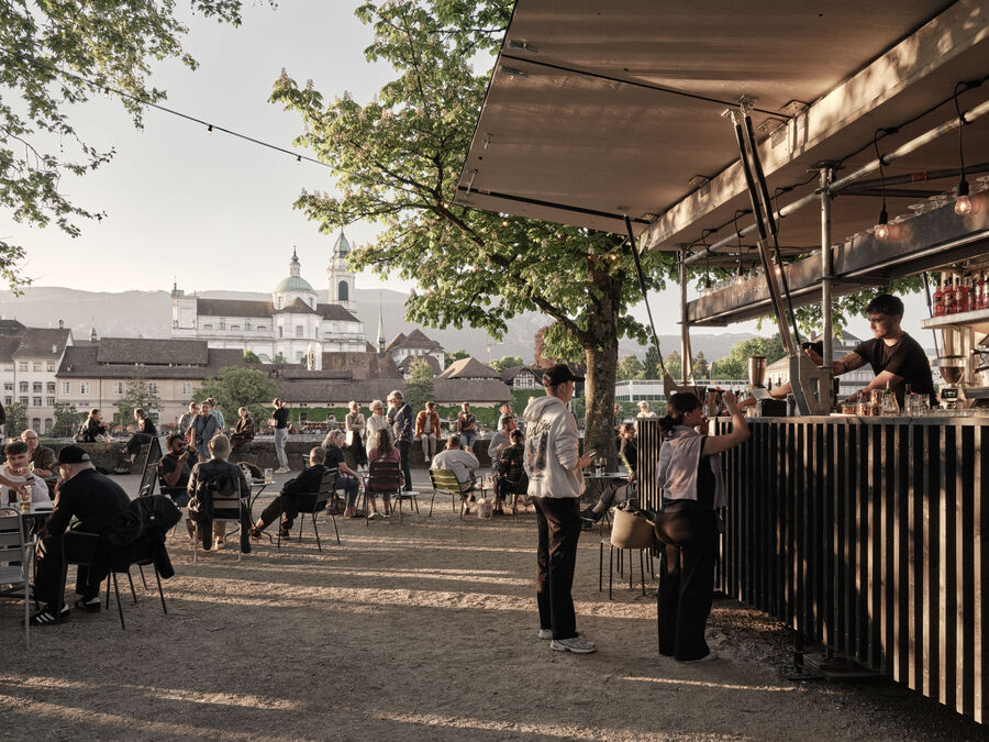 Architekturfotografie: Menschen sitzen und stehen bei der Buvette volAare am Kreuzackerplatz in Solothurn, einer temporären modularen Gastronomie am Aareufer.