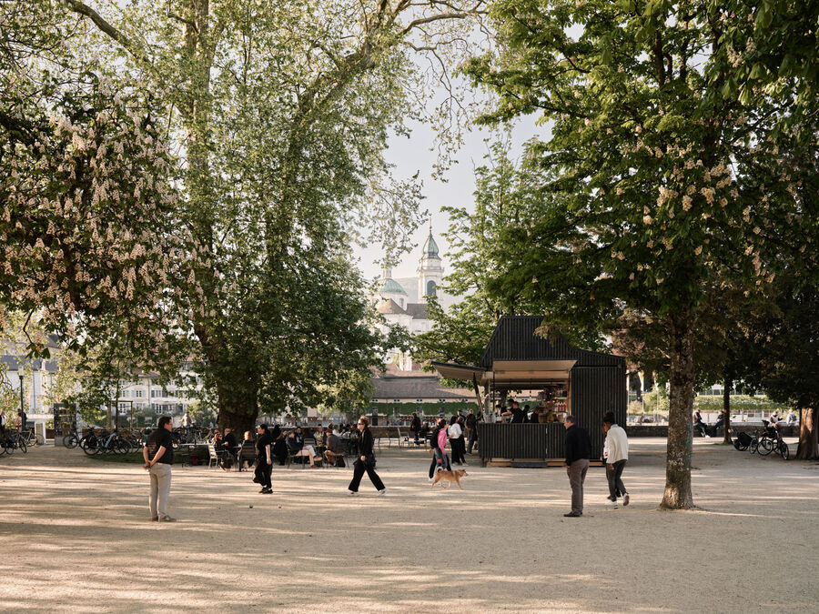 Architekturfotografie: Menschen sitzen und stehen bei der Buvette volAare am Kreuzackerplatz in Solothurn, einer temporären modularen Gastronomie am Aareufer.