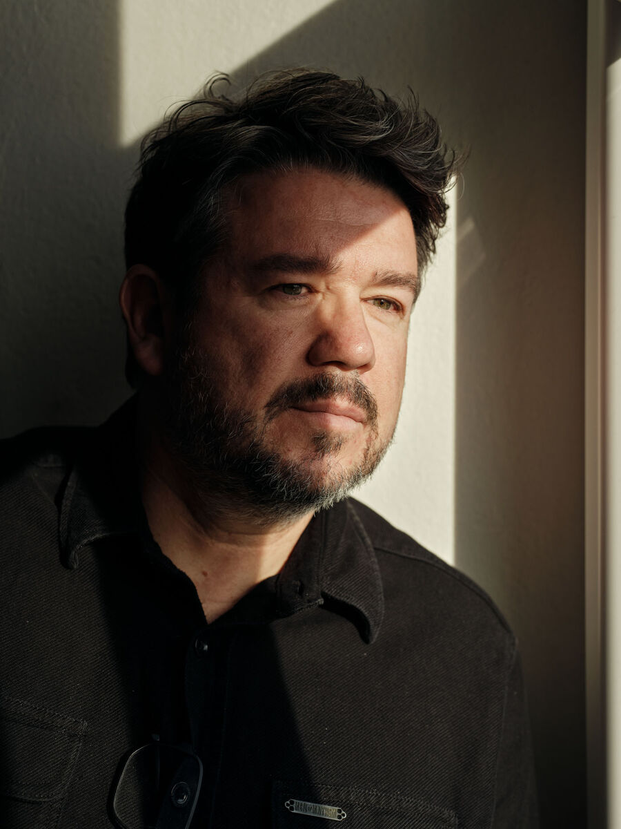 Nahportrait von Boris Szélpal im Seitenlicht, ruhiger Blick, reduzierte Atmosphäre im Atelier. Boris Szélpal sitzend an einem Tisch vor einem grossformatigen Gemälde in seinem Atelier.